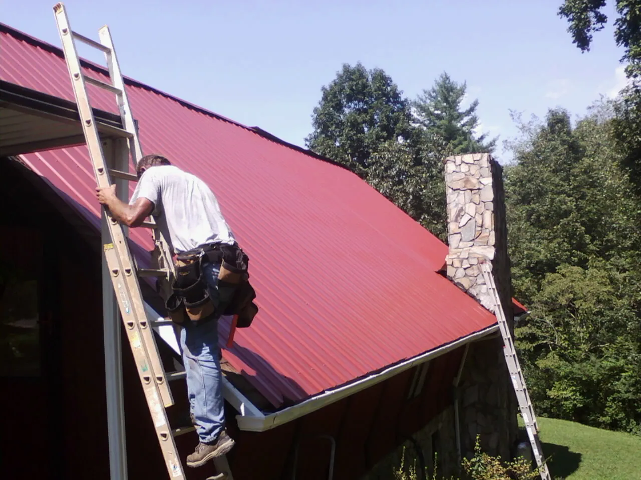 Roofer on a ladder beside a red metal roof and stone chimney — Soffit & Fascia Repair by Roofing Brothers in Tornado